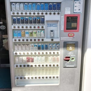 Puede incluir: Una máquina expendedora de tabaco gris con la palabra "TOBACCO" en letras azules en la parte superior. La máquina muestra varias marcas de cigarrillos y sus precios. Tiene una ranura de pago y un área de dispensación de productos.