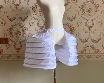 May include: A white pannier skirt support garment featuring a structured, multi-tiered design with horizontal boning channels. The piece is constructed from lightweight fabric and includes an adjustable waistband, designed to create volume at the hips.