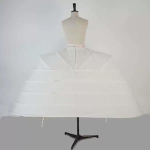 May include: A white bridal petticoat with multiple hoops, displayed on a dress form. The petticoat is designed to give volume to a wedding dress. The dress form is beige, and the stand is black.