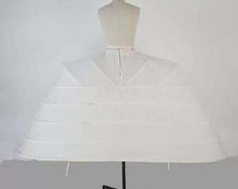 May include: A white bridal petticoat with multiple hoops, displayed on a dress form. The petticoat is designed to give volume to a wedding dress. The dress form is beige, and the stand is black.