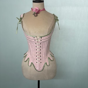 Puede incluir: Un corsé rosa con ribete verde y cordones blancos se exhibe en un maniquí. El corsé tiene un dobladillo festoneado y está adornado con correas de cinta verde. Un collarín de cinta rosa con una rosa y un colgante está alrededor del cuello del maniquí.