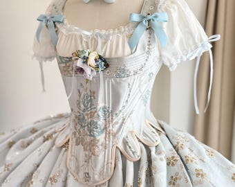 以下が含まれることがあります： 花柄の刺繍と金の装飾が施された水色のボールガウン。コルセット風のボディス、青いリボン付きの半袖、お揃いのチョーカーとリボンが特徴です。ボディスには花のブローチが付いています。