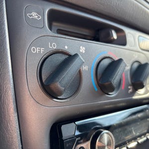 May include: Close-up of a car's dashboard featuring climate control knobs and a radio. The knobs are black and positioned on a gray panel. The temperature control dial has markings for OFF, LO, and HI, with blue and red indicators.