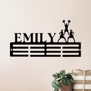 以下が含まれることがあります： 「EMILY」の文字と3本の水平バーが特徴の黒い金属製の壁飾り。チアリーダーのシルエットがバーの上に配置されています。メダルやリボンを飾るように設計されています。
