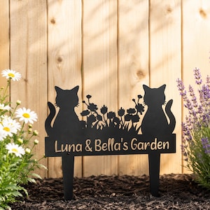 May include: A black metal garden stake featuring silhouettes of two cats sitting on either side of a floral arrangement. The rectangular base displays the text "Luna & Bella's Garden" in a light-coloured, sans-serif font, supported by two stakes.