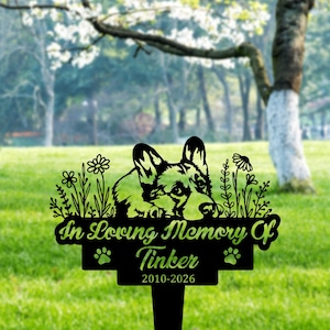 Może przedstawiać: Czarny metalowy palik pamiątkowy do ogrodu z motywem psa Corgi i napisem "In Loving Memory Of Tinker 2010-2026". Palik zawiera odciski łap i elementy kwiatowe.