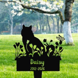 May include: Black metal garden stake with a dog silhouette, flowers, and a butterfly. The name "Daisy" and the years "2012-2026" are displayed. The background shows a green lawn and trees, typical of a garden setting.