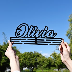 May include: Black metal sign with the name "Olivia" in a cursive font. The name is above a horizontal bar with three parallel lines. The sign is held up against a blue sky with trees in the background.