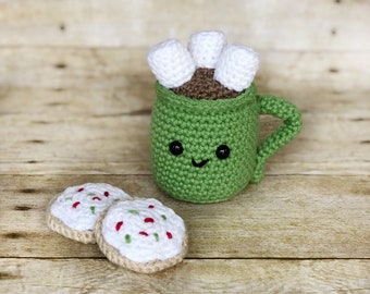 May include: A green crocheted mug with a smiling face, filled with brown crocheted hot chocolate and topped with white crocheted marshmallows. Two white crocheted cookies with red and green sprinkles are in front of the mug.