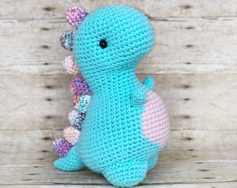 May include: A crocheted turquoise dinosaur toy with pink and purple accents on its back. The dinosaur has a pink belly and black eyes.