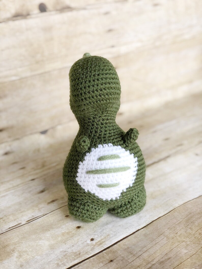May include: Green crocheted dinosaur toy with a white crocheted patch on its back.