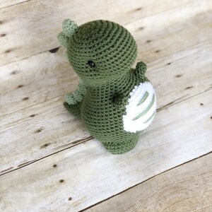 May include: A green crocheted dinosaur toy with white stripes on its back. The dinosaur has a small, round head with black eyes and a small, pointed nose.