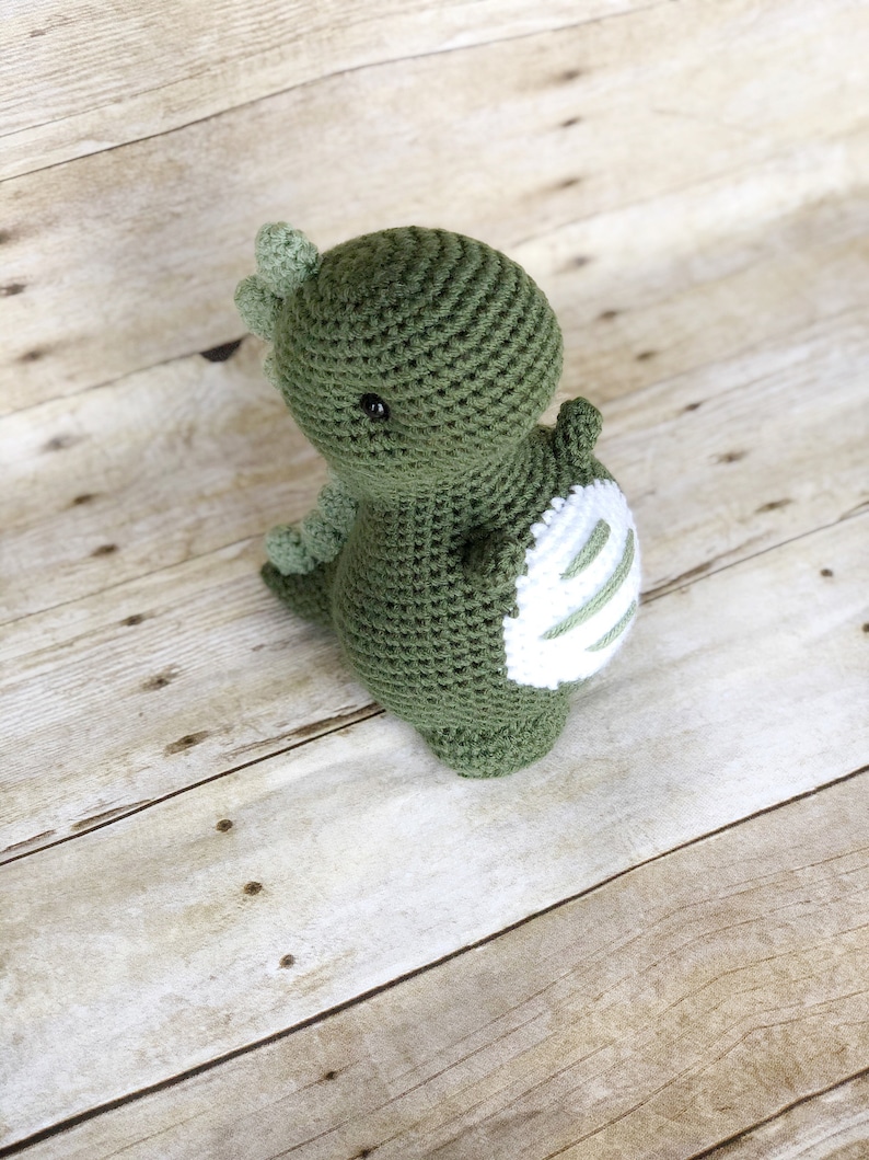 May include: A green crocheted dinosaur toy with white stripes on its back. The dinosaur has a small, round head with black eyes and a small, pointed nose.