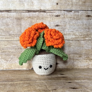May include: A crocheted flower pot with orange flowers and green leaves. The pot is white with a smiling face. The flowers are a vibrant orange color, and the leaves are green.