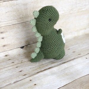 May include: Green crocheted dinosaur toy with a white patch on its back. The dinosaur has a green spiky back and black eyes.