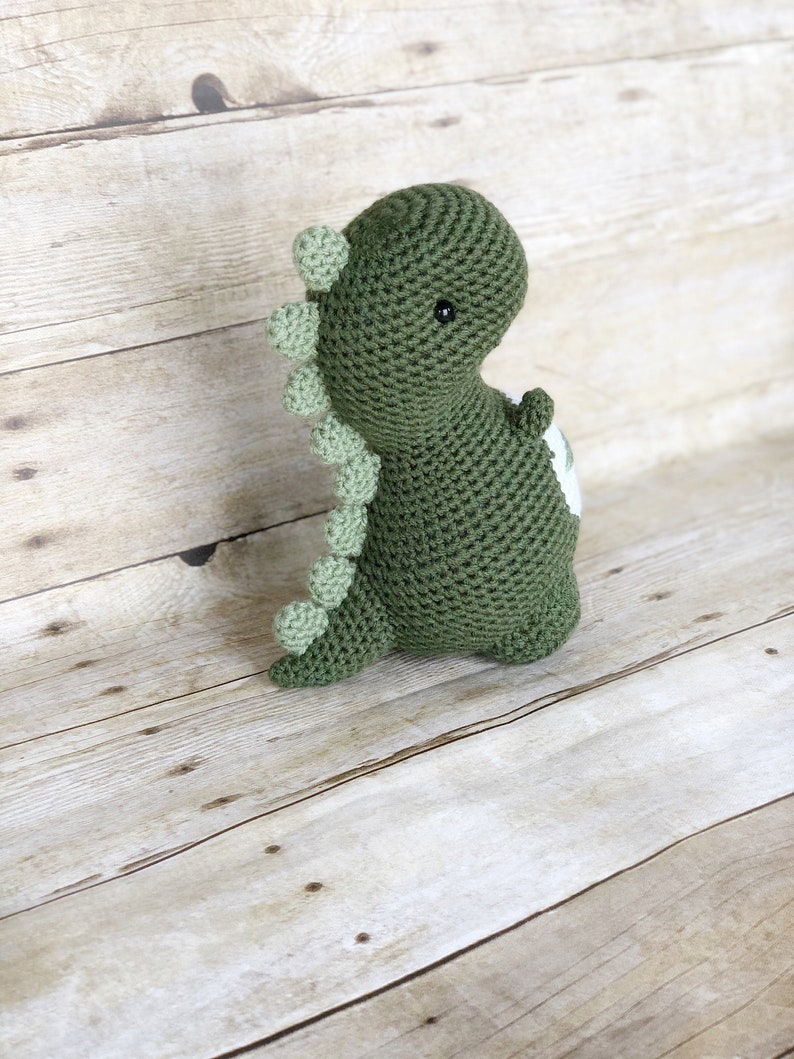 May include: Green crocheted dinosaur toy with a white patch on its back. The dinosaur has a green spiky back and black eyes.