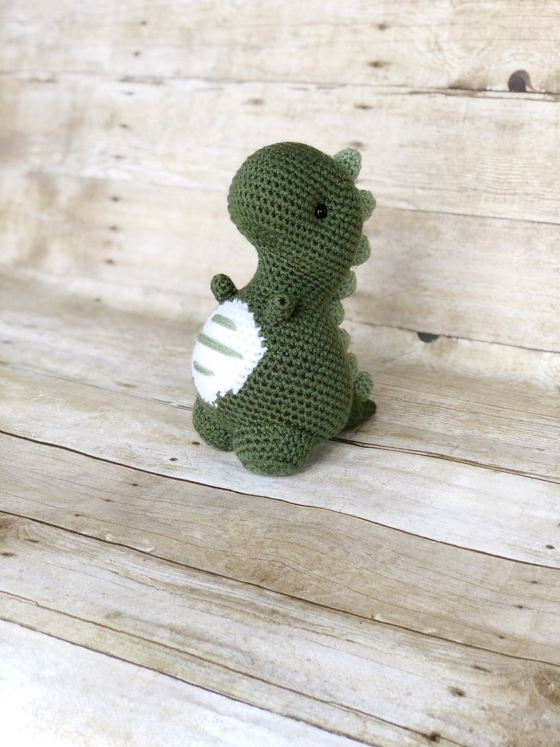 May include: A green crocheted dinosaur toy with white stripes on its belly. The dinosaur is standing on a wooden surface.