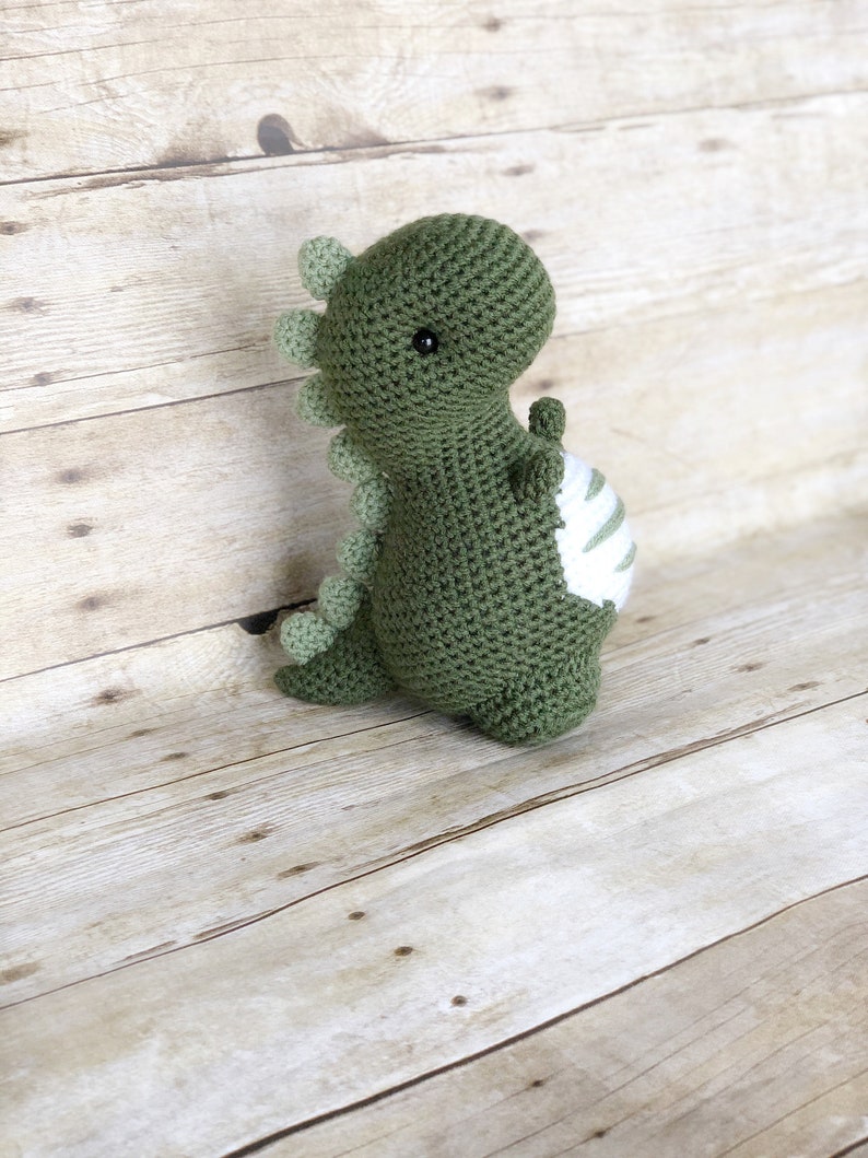 May include: A green crocheted dinosaur toy with white stripes on its belly. The dinosaur has black eyes and a small, pointed snout.