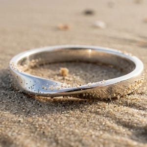 Puede incluir: Un anillo plateado con un diseño retorcido descansa sobre una playa arenosa. El anillo refleja la luz del sol, creando una apariencia metálica brillante. La arena es de color beige claro, con el océano al fondo. Es una pieza de joyería.
