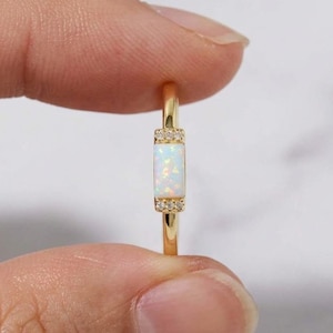May include: A gold ring with a rectangular opal gemstone. The opal is set vertically, with small, sparkling accent stones on either side. The ring's band is a thin, polished gold.