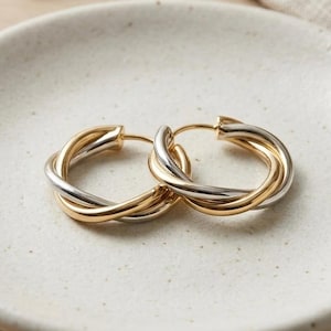 May include: A pair of hoop earrings featuring an intertwined design with alternating gold and silver-toned metal bands. The earrings have a polished, reflective finish and a circular shape, showcasing a mixed-metal aesthetic that combines two distinct colours.