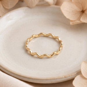 Puede incluir: Un anillo fino de color dorado que presenta un diseño ondulado o serpentino continuo. La banda tiene un acabado liso y pulido con un grosor constante a lo largo de toda su forma circular, creando una estética delicada y sencilla.