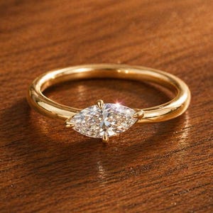 May include: A gold ring featuring a marquise-cut diamond. The diamond is set horizontally and catches the light. The ring has a polished gold band. The ring is presented on a wooden surface.