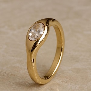 Peut inclure: Une bague en or dotée d'un anneau lisse et poli à la forme organique et fluide. La bague est sertie d'une unique pierre précieuse transparente de forme ovale placée sur le dessus, créant une transition continue entre le métal et la pierre.