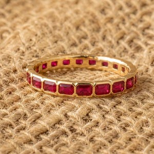 Peut inclure: Une alliance en or ornée d'une rangée continue de pierres précieuses rouges rectangulaires serties en rail. La bague présente un agencement uniforme de pierres sur toute sa circonférence, mettant en valeur une finition métallique polie.