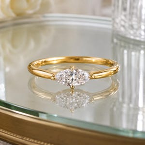 Op de afbeelding: Een gouden ring met een enkele, in marquise geslepen, heldere edelsteen in het midden. De ringband is glad en gepolijst met een dun, afgerond profiel. De edelsteen wordt op zijn plaats gehouden door kleine pootjes en heeft facetten.