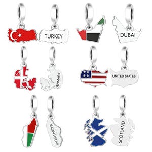 May include: A collection of silver-toned metal charms shaped like maps of Turkey, Dubai, Denmark, the United States, Madagascar, and Scotland. Each charm features a colored flag design on one side and the country name printed on the reverse side.