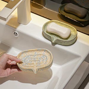 May include: An oval ceramic soap dish with a textured, speckled yellow and blue finish featuring a raised concentric oval pattern and a small drainage spout. The dish has a shallow basin and a rough, organic edge, designed for holding bar soap.