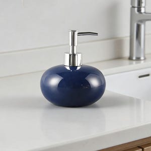 Può includere: Un dispenser di sapone in ceramica blu scuro di forma ovale con una finitura liscia e lucida. Il dispenser è dotato di un meccanismo a pompa color argento metallizzato sulla parte superiore, progettato per erogare sapone liquido o lozione.