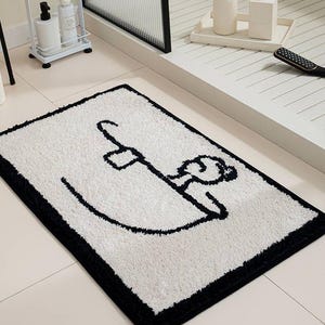 May include: A rectangular bathroom rug with a white shaggy texture and a black border. The centre features a minimalist black line drawing of a person relaxing in a bathtub, depicted with simple strokes showing a head, shoulders, and arm.
