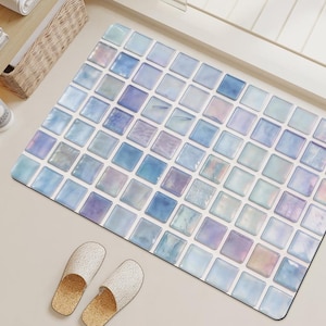 Op de afbeelding: Een rechthoekige badmat met een raster van vierkante tegels in diverse tinten lichtblauw, lavendel en wit met een glanzende, iriserende afwerking. Een paar beige, gestructureerde slippers liggen bij de linkerbenedenhoek van de mat.
