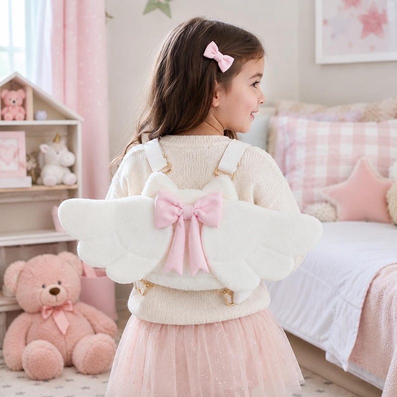 May include: A white plush backpack shaped like angel wings, with a pink bow in the center. The backpack has white straps and gold-colored clasps. The child is wearing a cream-colored sweater and a pink tulle skirt.