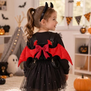 May include: A child wearing a black outfit with a red bow and bat wings attached to the back. The child is also wearing a black headband with horns. The outfit is a Halloween costume.