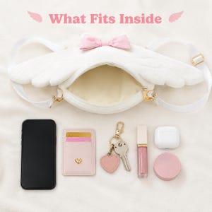 May include: White plush crossbody bag shaped like angel wings, with a pink bow and gold hardware. The bag is open, revealing its interior. Items inside include a phone, cardholder, keys, lip gloss, earbuds, and a compact.