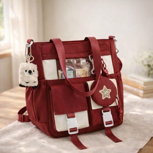 Puede incluir: Un bolso tote burdeos con detalles en crema, múltiples bolsillos y cierres de hebilla blancos. Se adjunta un pequeño monedero redondo con un diseño de estrella y un llavero de peluche. La bolsa tiene un bolsillo transparente con objetos pequeños.