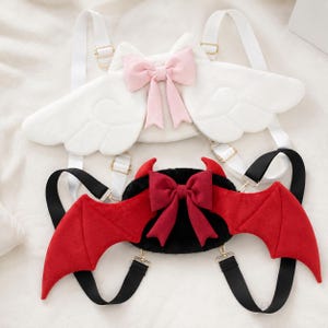 May include: Two novelty backpacks, one white with angel wings and a pink bow, the other black with red devil wings and a red bow. Both have adjustable straps and gold-colored hardware.