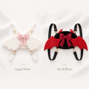 May include: Two novelty backpacks, one white with angel wings and a pink bow, the other black with devil horns and red wings. Both have adjustable straps and gold-colored hardware. The text reads "Angel White" and "Devil Black".