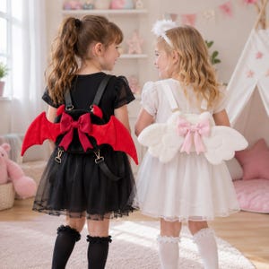 May include: Two young girls wearing themed backpacks. One backpack is black with red bat wings and a red bow. The other is white with angel wings and a pink bow. Both girls are wearing dresses and knee-high socks.