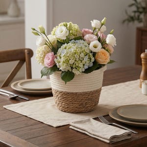 May include: A floral display in a two-tone woven basket. The basket has a white rope top and a brown woven base. The flowers include white, pink, and peach roses, and green and blue hydrangeas. The arrangement is on a wooden table with place settings.