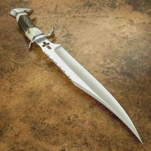 May include: A large, silver-colored knife with a serrated edge and a cross-shaped cutout near the handle. The handle is made of a light-colored material with dark brown accents. The knife has a curved blade and a pointed tip.