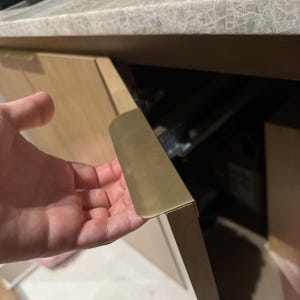 Könnte beinhalten: Nahaufnahme einer Schranktür mit einem messingfarbenen Griff. Der Griff ist rechteckig und an einem hellen Holzschrank befestigt. Der Schrank ist teilweise geöffnet und gibt den Blick ins Innere frei.