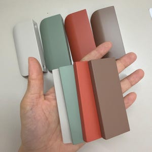 May include: A collection of eight rectangular cabinet pulls in four distinct matte colors: off-white, sage green, terracotta orange, and taupe brown. Each pull features a smooth, curved edge design with visible screw holes on the mounting tabs.
