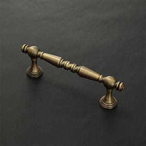 Op de afbeelding: Een meubelgreep met een antieke messing afwerking. Het beslag is voorzien van een centrale cilindrische stang met decoratieve geribbelde details en twee afgeronde eindkappen, ondersteund door twee ronde voetstukken die vanaf de basis uitsteken.