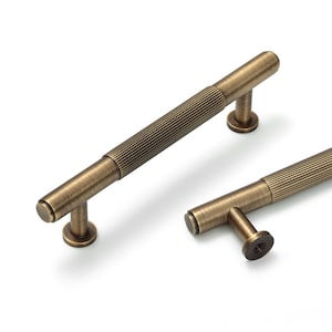 May include: Two cylindrical cabinet pulls with a brushed antique brass finish. Each handle features a central section with a vertical ribbed texture and smooth ends. The pulls are mounted on two circular bases, with one base showing a screw hole.