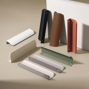 May include: A selection of contemporary cabinet pulls in diverse colours: white, beige, navy, brown, terracotta, light grey, and sage green. These metal pulls feature a minimalist design with a curved handle. Measurements are in centimetres.