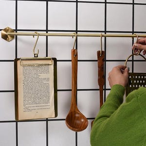 May include: A brass-colored kitchen rail with hanging utensils and a clipboard. The utensils include a wooden spoon, a spatula, and a grater. The clipboard holds a page of text. The background is a white tile wall with black grout.